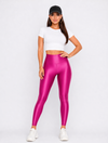A woman modeling shiny metallic magenta-pink high-waisted leggings, paired with a white crop top, black baseball cap, and white sneakers, against a light gray studio background.