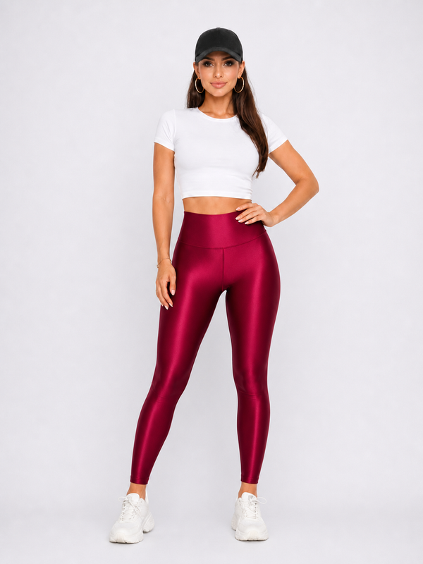 A woman modeling shiny burgundy high-waisted leggings, a white crop top, and white sneakers against a light gray studio background.