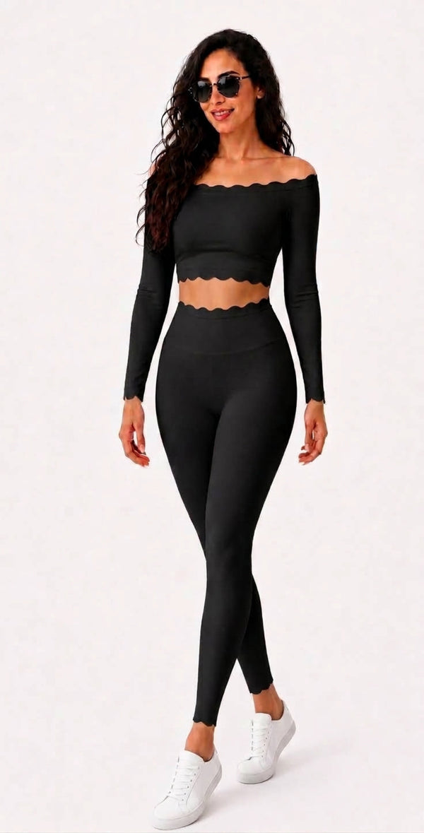 A woman modeling a black two-piece activewear set consisting of a long-sleeve off-the-shoulder crop top with a scalloped edge and matching high-waisted leggings, paired with white sneakers against a plain white background.
