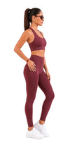 A woman modeling a matching burgundy activewear set consisting of a sports bra and high-waisted leggings, paired with white sneakers, against a plain white background.