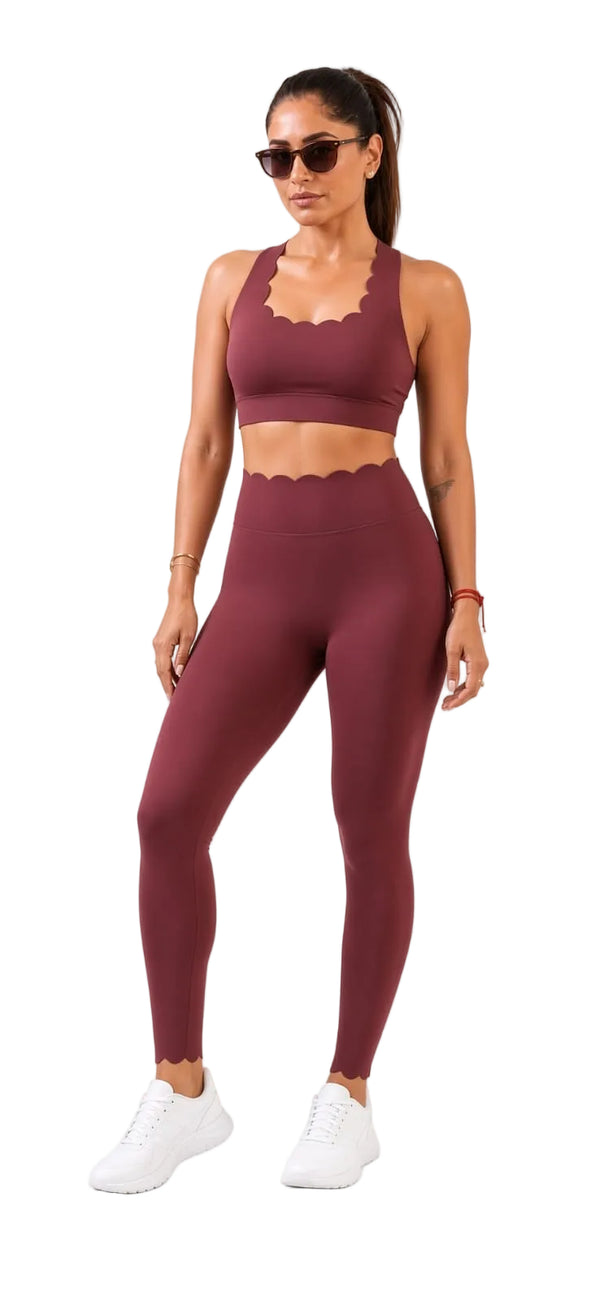 A woman modeling a matching burgundy activewear set consisting of a scoop-neck sports bra with a wavy scalloped neckline and high-waisted leggings, paired with white sneakers and sunglasses against a white background.