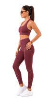A woman modeling a matching burgundy activewear set consisting of a sports bra and high-waisted leggings, paired with white sneakers, against a plain white background.