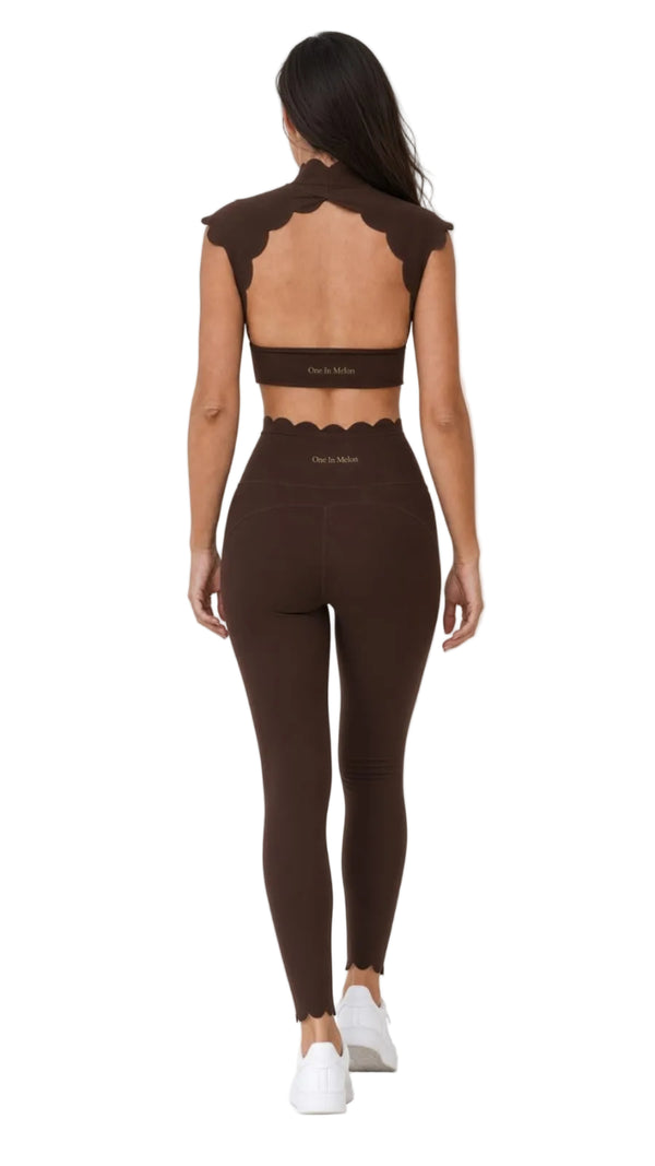 A woman shown from the back wearing a matching dark brown activewear set consisting of a scalloped open-back crop top and high-waisted leggings, paired with white sneakers against a bright white studio background.
