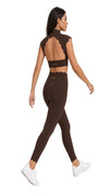 A woman modeling a matching dark brown activewear set consisting of a scalloped back sports bra and high-waisted leggings, shown from a rear three-quarter view against a bright white studio background.