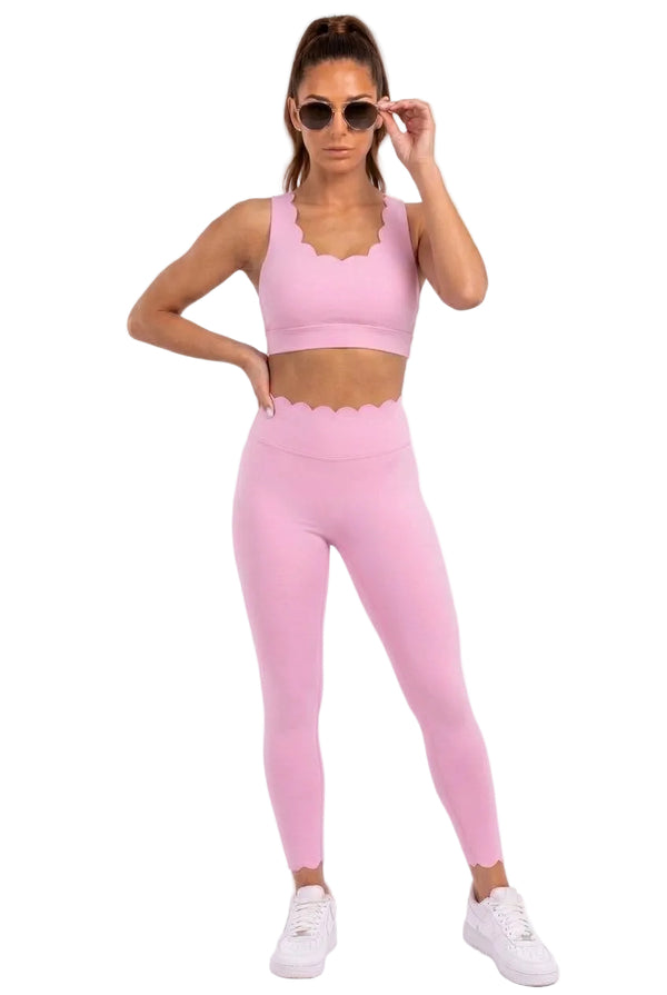 A woman modeling a matching light pink activewear set consisting of a scoop-neck sports bra and high-waisted leggings with a scalloped edge, paired with white sneakers and sunglasses against a white studio background.